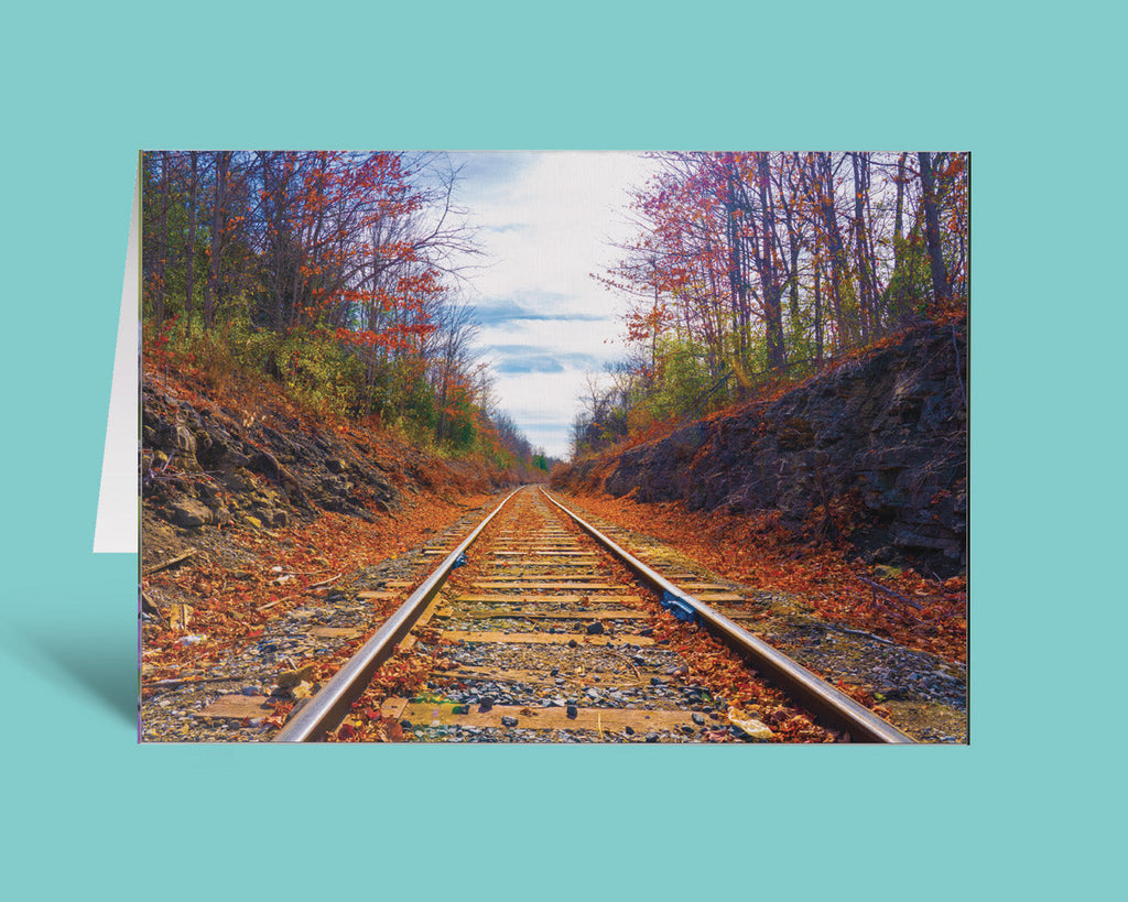A vertical greeting card featuring a photograph of railroad tracks running through an autumn forest with fallen leaves, shown standing against a solid turquoise background.