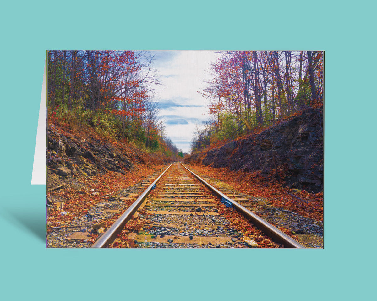 A vertical greeting card featuring a photograph of railroad tracks running through an autumn forest with fallen leaves, shown standing against a solid turquoise background.