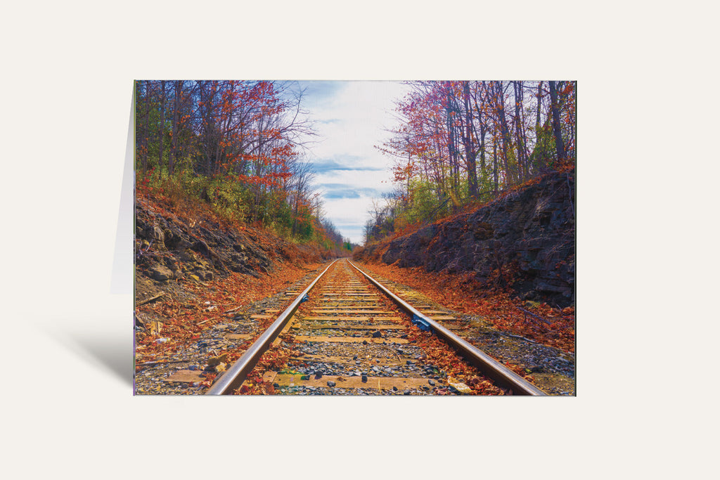 A vertical greeting card featuring a scenic photograph of railroad tracks running through an autumn forest with orange and red leaves, set against a plain off-white background.