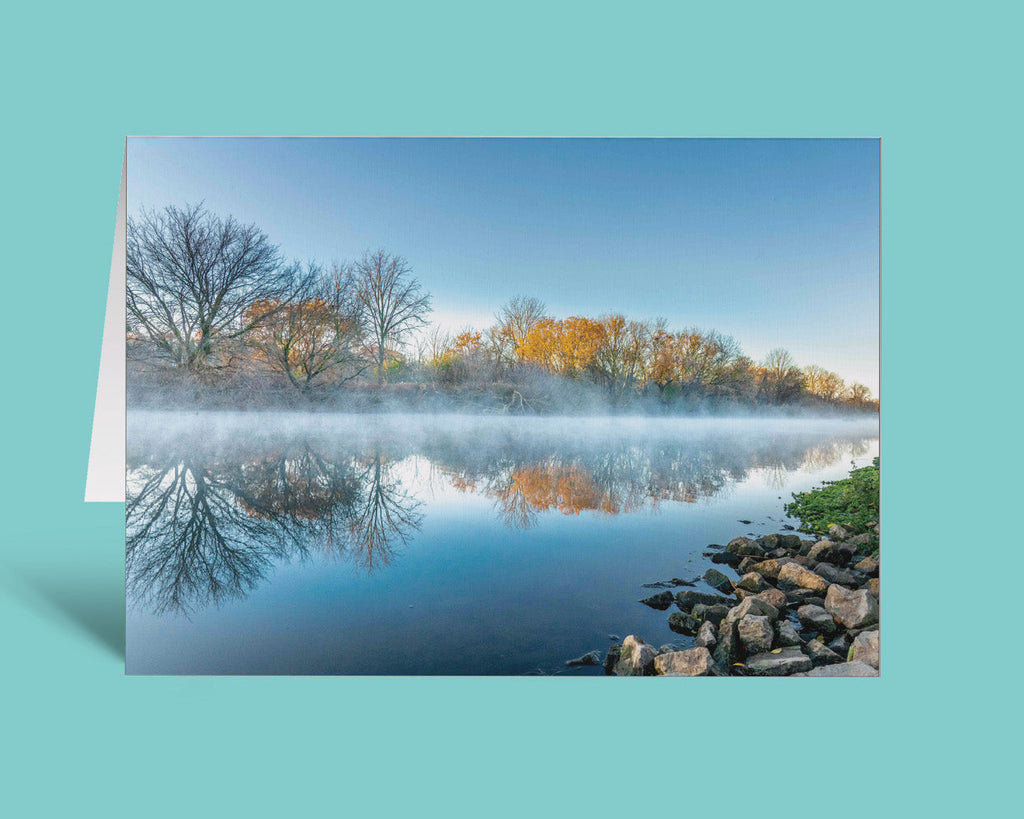 A folded greeting card featuring a scenic landscape photograph of a calm river with mist, bare trees, yellow autumn foliage, and rocks along the shore, displayed against a solid teal background.
